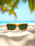 Eagle Beach Gold sunglasses resting on bright sand with clear blue ocean and palm trees in the background.