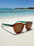 Wariruri Aura sunglasses – Green Frame, Amber Lens resting on white sandy beach near turquoise water.